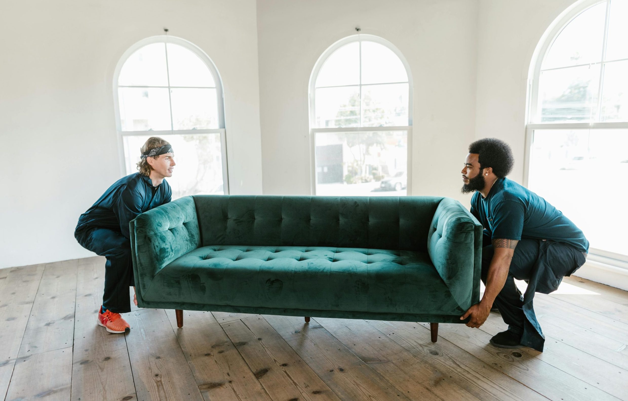 Two men carrying a green sofa in a sunlit room with wooden floors and arched windows.